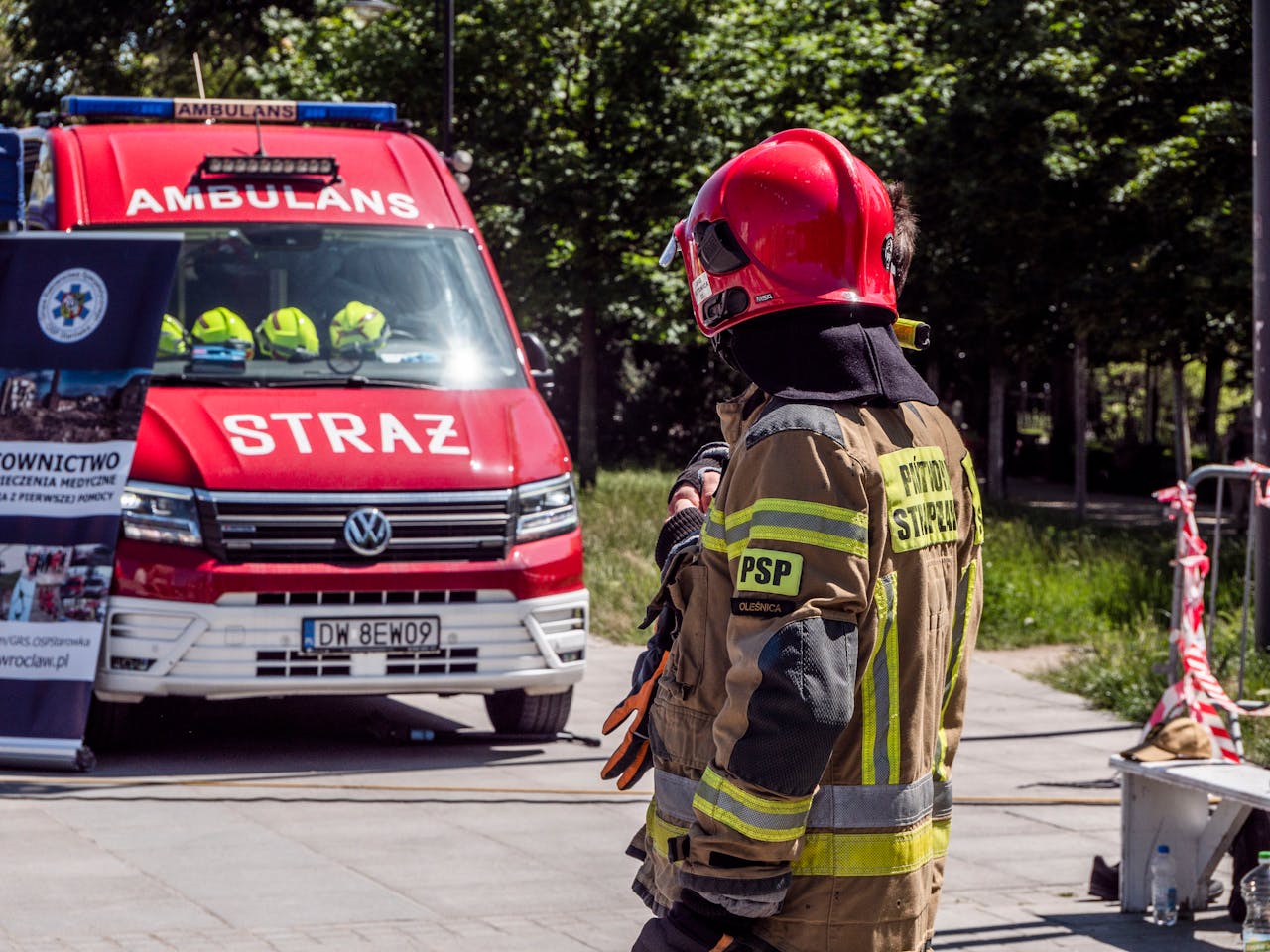 Firefighter in action near red ambulance labeled 'STRAŻ' outdoors in a daytime emergency scene.